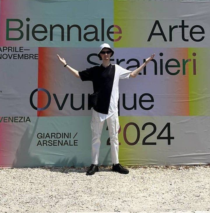 Антон с раскинутыми руками у баннера Biennale Arte 2024 «Stranieri Ovunque»
