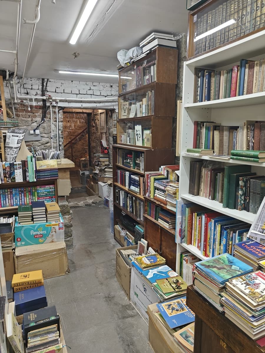 Подвальный зал: книги на стеллажах, на полу и в коробках, каменная лестница в глубине
